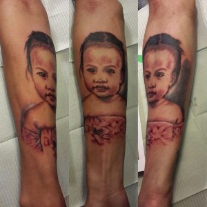 A forearm tattoo in black and gray shows a realistic portrait of a young child with short hair and a floral-patterned outfit, viewed from different angles.