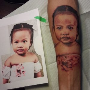 A forearm tattoo of a young girl is shown next to the reference photo. The tattoo and photo both show the girl with braided hair, wearing a white dress with a pink flower design.