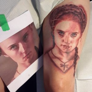A realistic tattoo of a young woman with braided hair and a necklace is shown on a persons arm, next to the reference photo the tattoo is based on. The images closely resemble each other.