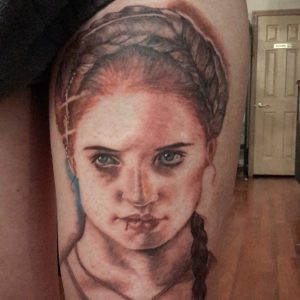 A realistic tattoo of a young woman with braided hair and piercing blue eyes is inked on someones upper arm or thigh. The background shows a wooden floor and a closed beige door.