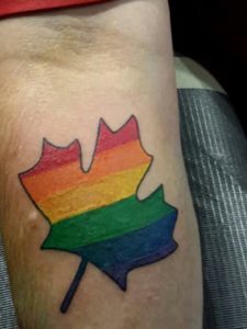 Arm displays a tattoo of a maple leaf filled with rainbow colors and outlined in black.