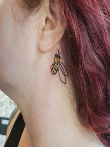 Tiny bee tattoo in black and yellow ink near a person's ear, with burgundy hair visible.