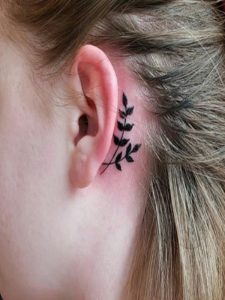 Botanical tattoo of a black sprig with leaves adorns the area behind a person's ear.