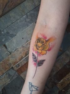 Yellow rose tattoo with faint red and orange watercolor effect on a person's forearm.