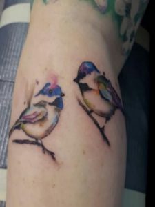Arm tattoo features two colorful birds perched on a branch in watercolor style.