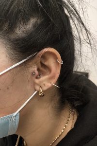 Person's ear with multiple piercings including helix, tragus, and lobe, accessorized with various hoop and stud earrings.