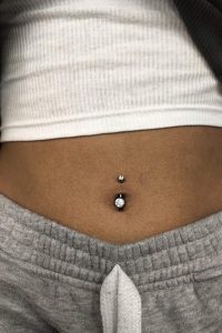 Close-up of a navel piercing with a large, clear gem, and a smaller gem above, on a person wearing a white tank top and grey sweatpants.