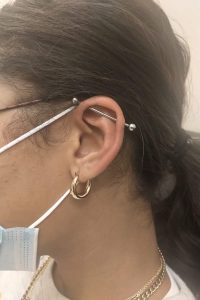 Person wears face mask and glasses, displaying an industrial piercing, and a golden hoop earring.