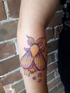 Arm displays a colorful moth tattoo with pink wings and small stars around it.