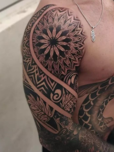 Shoulder and arm tattoo displays a mandala design, tribal patterns, and leaf elements in black ink.