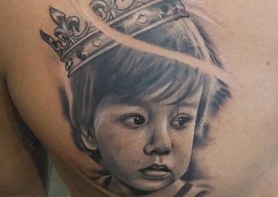 Chest tattoo of a child's portrait wearing a crown, rendered in detailed black and gray ink.
