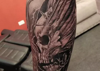 Tattoo on a forearm depicts a skull with wings, lightning bolts, and intricate shading in black and gray.