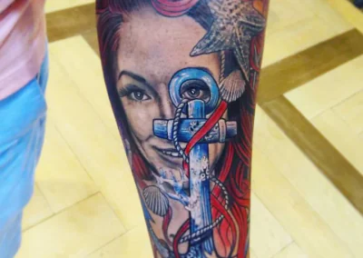 Forearm tattoo shows a woman's face with red hair, accented by a blue anchor, starfish, and seashells.