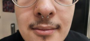 Person with nose piercings and a thin mustache wears glasses.