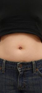 Close-up of a pierced navel with a jeweled barbell, set against blue denim jeans and a black shirt.