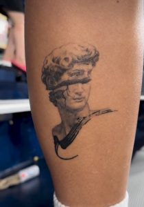 A tattoo of a bust with a crack across the face, on a person's arm.
