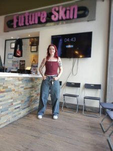 Red-haired woman with tattoos stands smiling in front of Future Skin sign and three chairs.