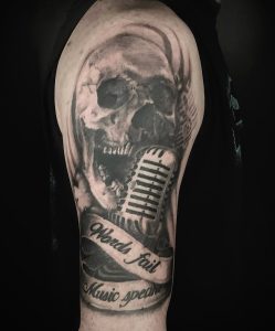 Upper arm tattoo featuring a skull, vintage microphone, and banner with the text 'Words fail, Music speaks'.