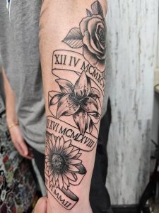 Forearm tattoo featuring a rose, lily, and sunflower with banners displaying Roman numerals.