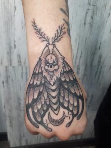 Hand shows a moth tattoo with a skull design and crescent moon detail.
