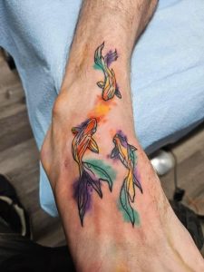 Foot shows a tattoo of three colorful fish with watercolor-style splotches in orange, green, and purple.