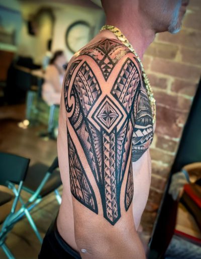 Man shows off a bold, black tribal tattoo design covering his shoulder and upper arm, wearing a thick gold chain.