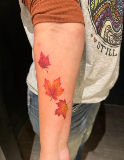 Forearm shows tattoo of three colorful autumn maple leaves in red, orange, and yellow.