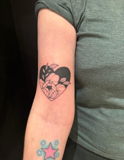 Arm shows a black heart tattoo filled with line art of flowers and leaves, plus a faded pink star tattoo.