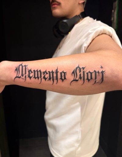 Person's arm with a 'Memento Mori' tattoo in black gothic script.