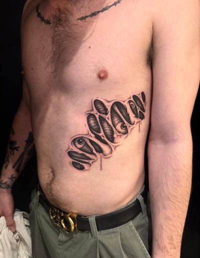 Man's torso shows a tattoo of eyes peeking through torn skin, with a barbed wire tattoo on his chest.