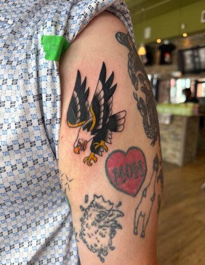 Arm with tattoos including an eagle, a heart that says "MOM", a chicken head, and another animal.