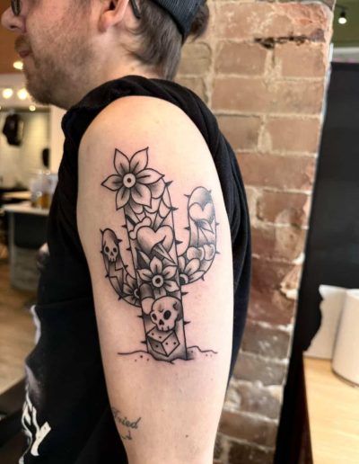 Person's arm with a black and white cactus tattoo featuring flowers, hearts, skulls, and a dice.
