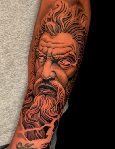 Forearm tattoo featuring a detailed portrait of Zeus with flowing hair and beard.