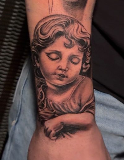Tattoo on forearm shows a grayscale image of a cherubic child with eyes closed and curly hair.