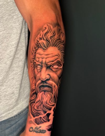 Arm displays a detailed black and gray tattoo of a bearded man's face with flowing hair and the text 'Adam'.