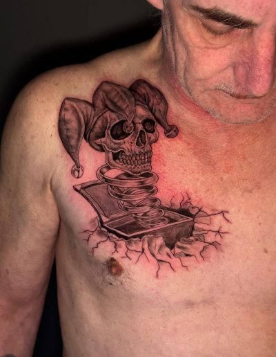 Man's chest features a tattoo of a jester skull popping out of a jack-in-the-box.