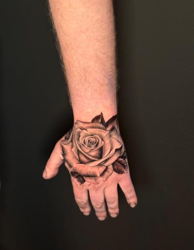 Hand displays a detailed rose tattoo with shading and leaves against a dark background.