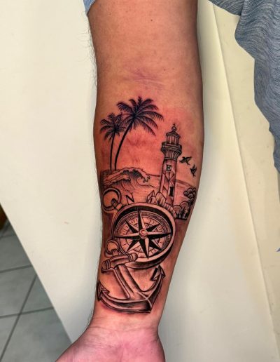 Nautical tattoo: compass, anchor, lighthouse, palm trees, and waves on an arm.