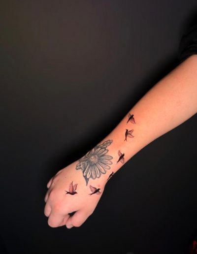 Tattoo of a daisy with flying birds on a person's arm and hand.