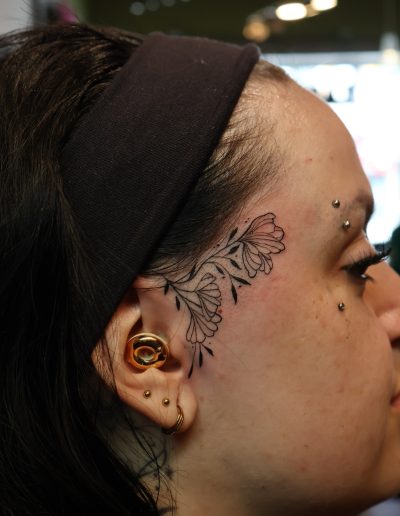 Close-up of a woman's face with a delicate floral tattoo on her temple and ear piercings.