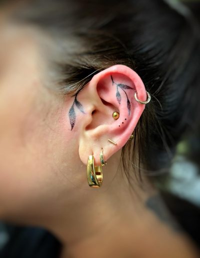 Close-up of a woman's ear with minimalist leaf tattoos and gold hoop earrings.