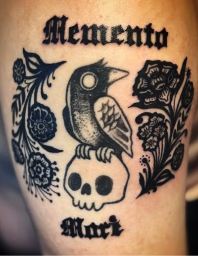 Black ink tattoo of a crow on a skull, with "Memento Mori" text and floral elements.