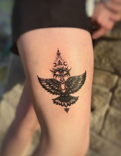 Black ink tattoo of an owl with an all-seeing eye and moon phases on a person's thigh
