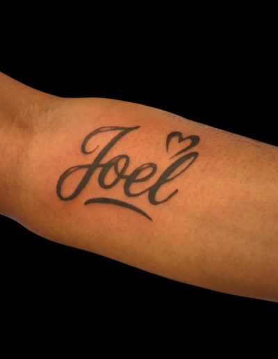 Close-up of a black ink tattoo of the name "Joel" with a small heart on a person's arm.