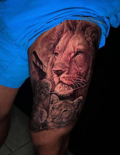 Realistic black and grey tattoo on an upper thigh featuring a male lion, lioness, two cubs, and a lion silhouette on a cliff.